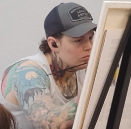 Eleena- Monk A focused artist with tattoos painting a portrait on an easel.